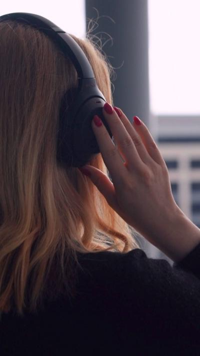 The back of a woman's head, with black headphones on. Her hand holds onto the right headphone, and has red nail varnish on. She has gingery/blonde, shoulder-length hair.