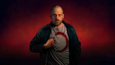 Actor Matthew Gurney wearing a T-shirt with a red unfinished circle and his bloodied hand on his chest, against red background.