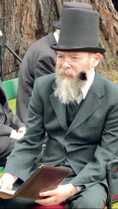A man with a black top hat and dark suit with a long beard. Her is sat on a chair reading a brown leatherbound book.