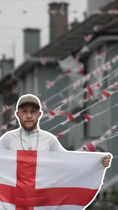 A wearing a baseball cap and gold chain holds up a Cross of St George, with terraced housing behind him.
