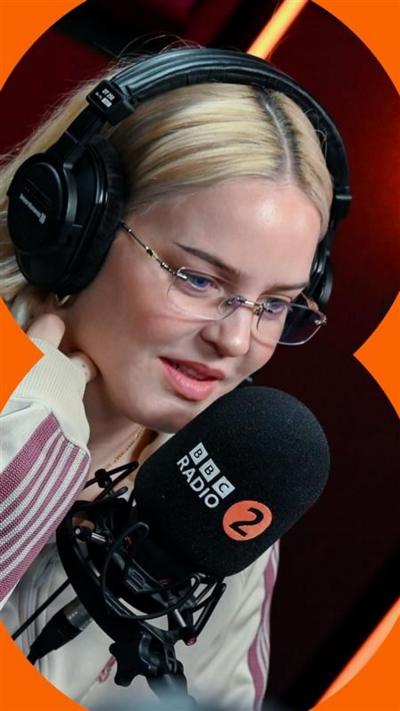 Anne-Marie sits in a Radio 2 studio - she has shoulder length blonde hair and wears a grey and red tracksuit
