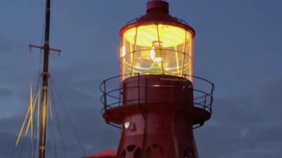 The top of a lighthouse is lit up at dusk. It's glowing with an orange light and a ship's mast can be seen to the left of it. The sky is cloudy and a dark blue-grey colour.