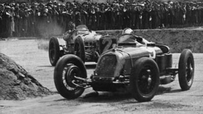 a black and white photo of two cars racing in the first Grand Prix race in Britain at Brooklands racetrack