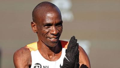 A head and shoulders shot of Eliud Kipchoge in a running vest and holding his gloved hands up in prayer.