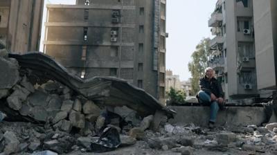 A man sits on a concrete, surrounded by rubble.