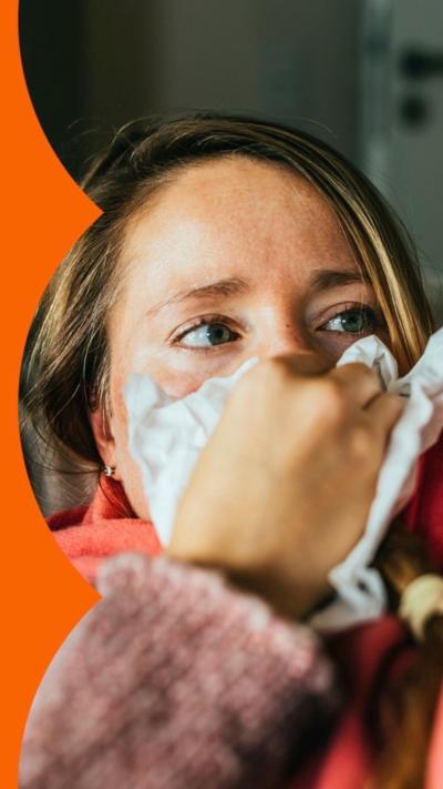 A woman on a sofa, holding a tissue up to her nose, wearing a red scarf.