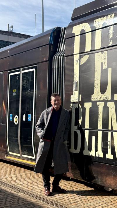 A man in a long grey coat stood next to a tram with the words Peaky Blinders emblazoned on the sides in cream lettering