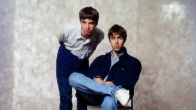 Noel Gallagher and Liam Gallagher from the band, Oasis, are in front of a beige background at a photoshoot in a hotel in Tokyo, September 1994. Noel is standing on the left and has brown hair, blue eyes, wears a white shirt, dark blue trousers, and is leaning over slightly. Liam is on the right, and has brown hair, blue eyes, wears a blue shirt, a dark blue jacket, blue jeans, white trainers, and is sitting down on a chair.