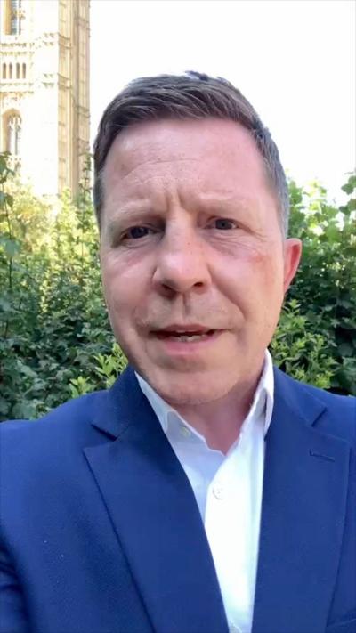 Male journalist wearing a blue suit with a white shirt, standing in front of a hedge