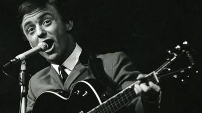 Gerry Marsden sings into a microphone while playing the guitar