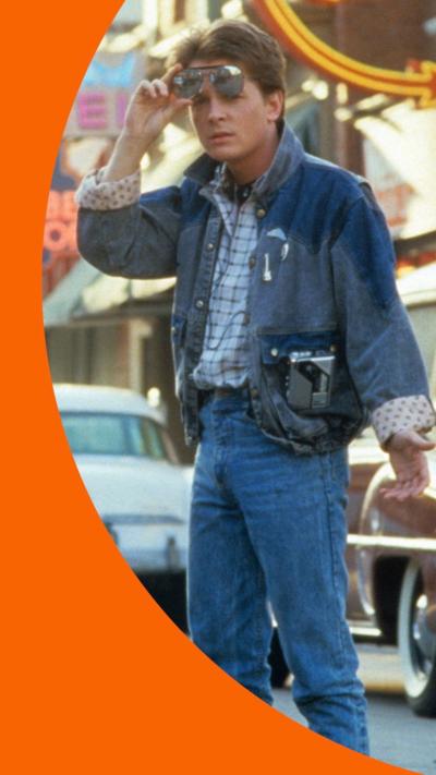 Michael J Fox as Marty McFly in Back to the Future. He wears a blue denim outfit and holds sunglasses above his eyes 