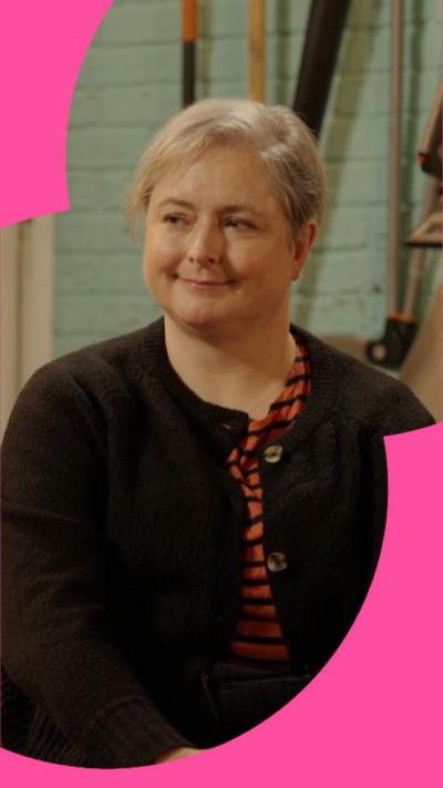 Siobhan McSweeney is sat smiling with her hands clasped on her knee. She's wearing a black cardigan and is looking to the side.