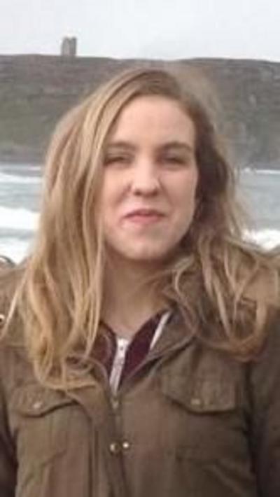 Natalie McNally standing on a beach with the sea visible behind her. She has long blonde hair and is wearing a dark green coat. She is smiling. 