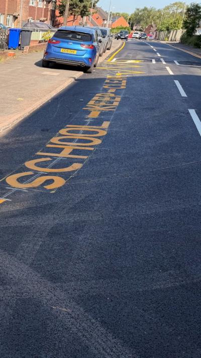 View of a road, on left hand side you can see yellow road markings saying, 'school-keep-clear'. A blue car is straddling the kerb on left hand side. 