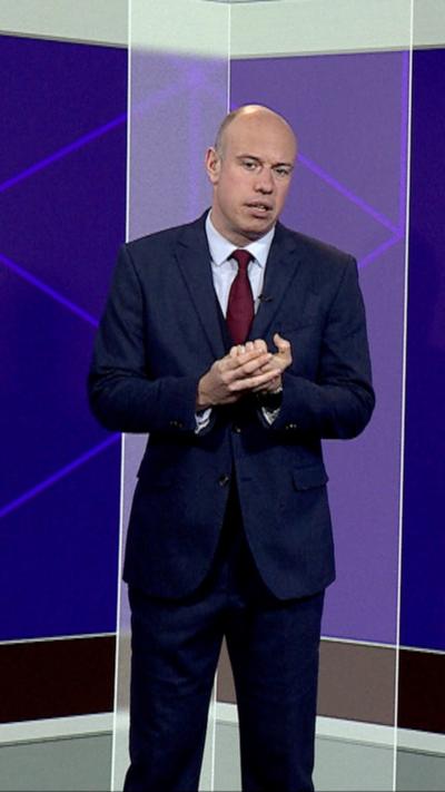 A man in a dark suit stands in front of a purple BBC studio background