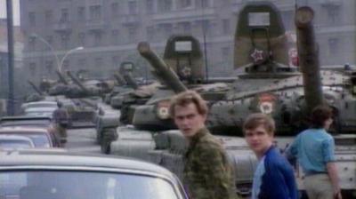 Pedestrians walk past cars and army tanks