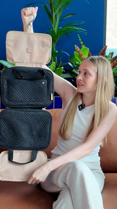 A young woman in a white top with long blonde hair, sitting on a pink sofa in front of a blue wall and green plants. She is holding up a toiletry caddy made up of several handbag-sized compartments.