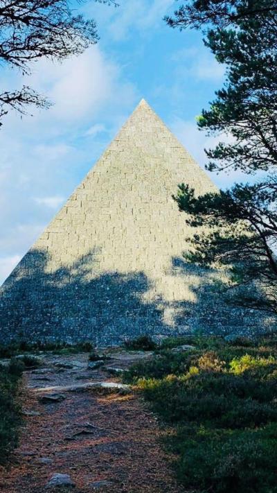 Pyramid-shape cairn at the edge of woodland, with the shadow of trees across the structure.