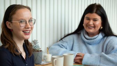 Two women sit either side of a table. They are both smiling at the camera. There are mugs and a plant between them