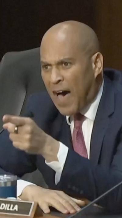 Senator Cory Booker points his finger and shouts toward the witness seat from a large dias in a US Senate hearing room.