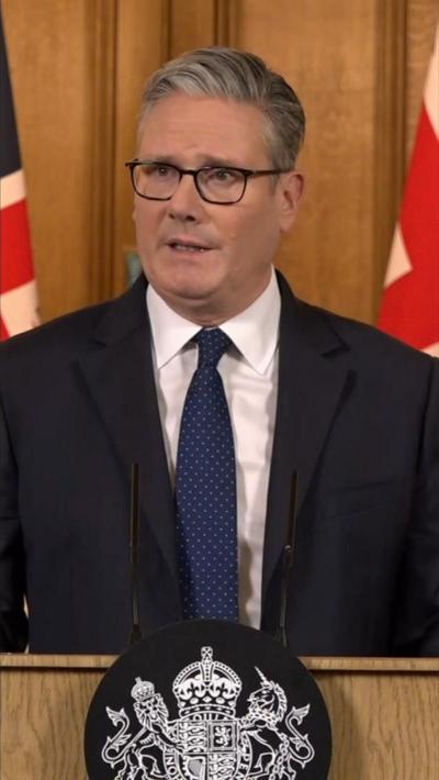 Sir Keir Starmer in a black suit and navy blue tie, stood at a podium