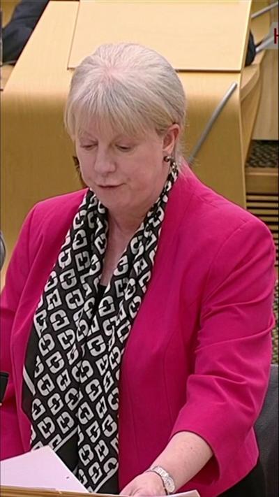 Woman with short blonde hair in a pink jacket and black and white scarf.