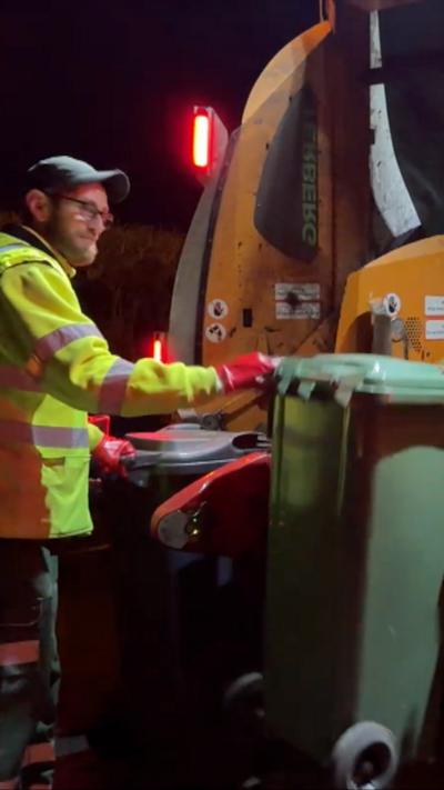 A male refuse collector in hi vis jacket and a cap at night emptying a bin onto the bin truck