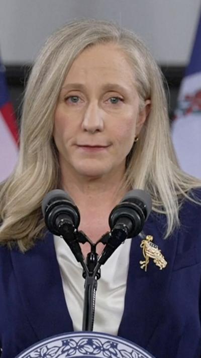 A woman with blonde hair standing in front of microphones, while wearing a dark blazer and white blouse