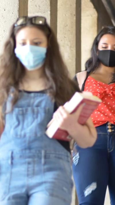 Two women wearing casual clothes and face masks and carrying books walk down a corridor