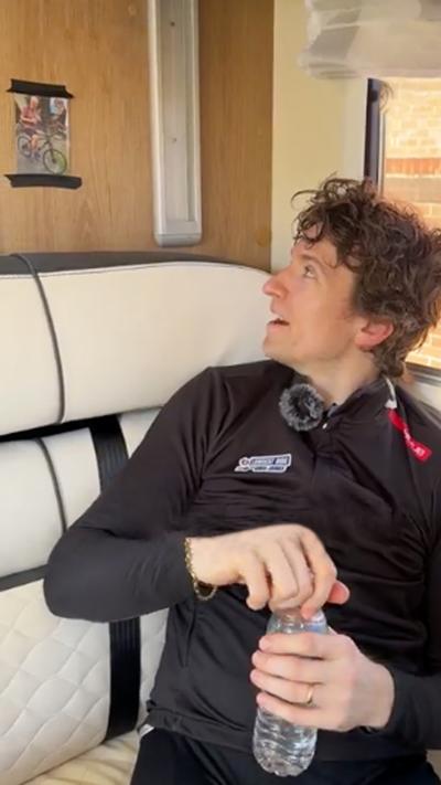 Greg James, wearing a black cycling top, is sat looking to the side of a picture of his dad which is stuck to the wall