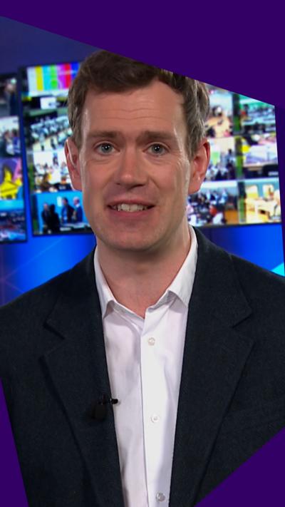 Phil Sim - man in shirt and suit, standing in front of a bank of TV monitors