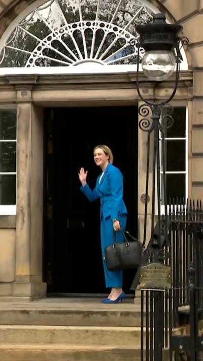 Jenny Gilruth is standing in the doorway of Bute House wearing a bright blue suit and waving.