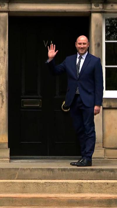 Neil Gray is smiling and waving in the doorway of Bute House.