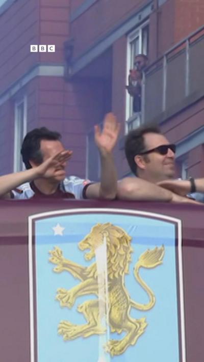 Two men on a claret coloured bus with a lion crest on a sky blue background