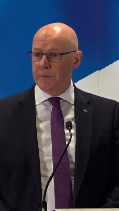 First Minister John Swinney is standing in front of a podium with a mic, with a saltire background.