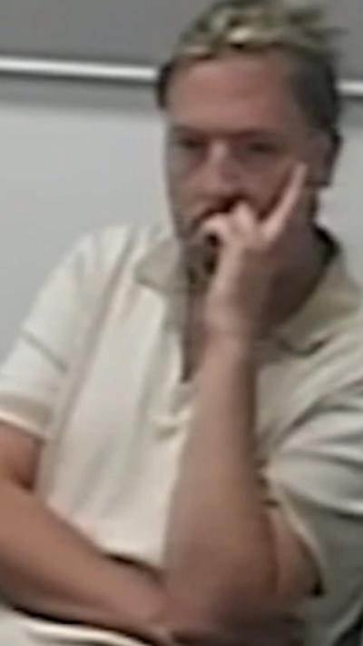 Jamie Varley in a police custody suite wearing a white polo shirt. He has longish blond hair