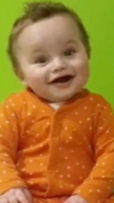 Baby Preston wearing an orange babygrow looking at the camera and smiling wiht a green background