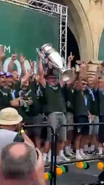 Players on a stage are holding a silver trophy high above their heads