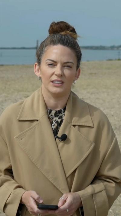 Female reporter with brown tied-up hair and brown coat stands on beach