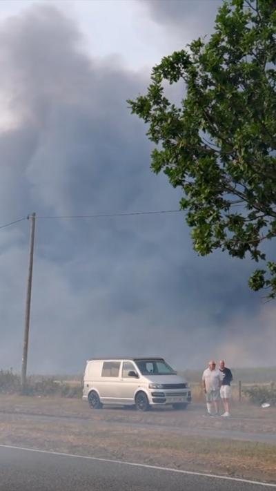 Plumes of black smoke drift over a field, there is a van and two people by the side of a road