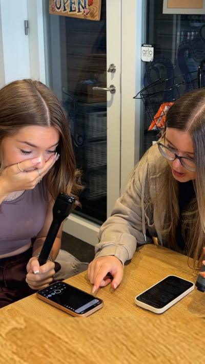 Two girls sitting at a table, one pointing at a phone screen and the other covers her mouth with her hand.