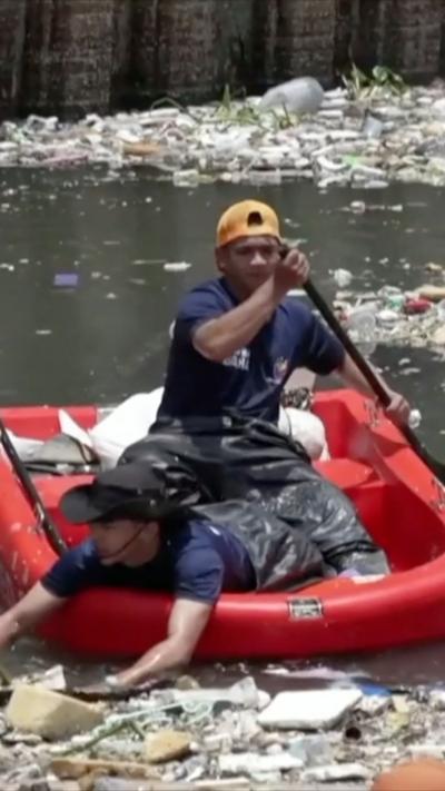 Two men row in a red boat surrounded by rubbish.