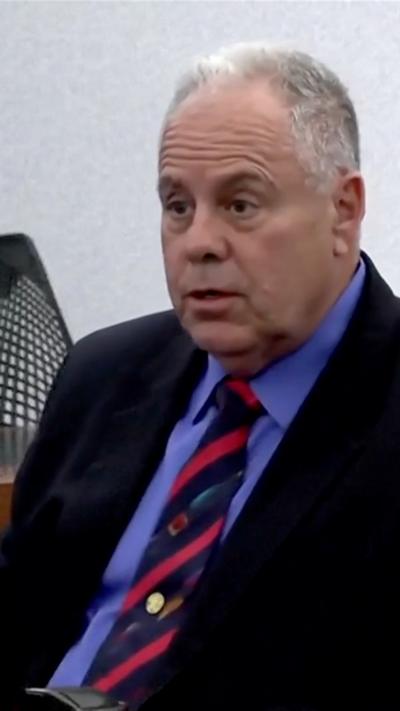 A man in a dark suit with grey/ white hair sitting.