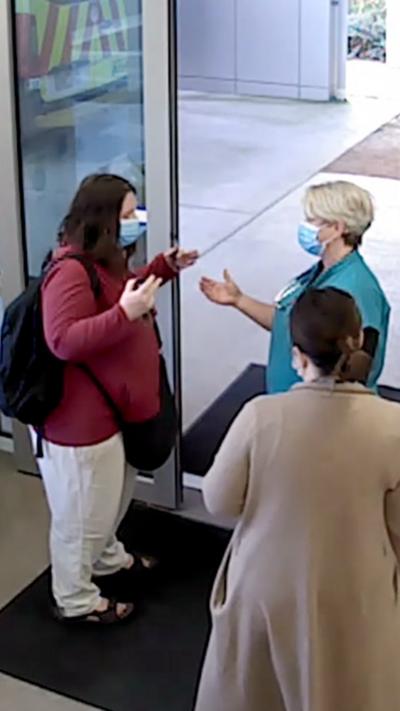 Three women stand in front of a sliding door talking with their hands raised while wearing blue face masks.