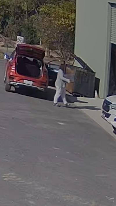 A person in a grey tracksuit holding an object walks into a shed while a red car is parked with its boot open.