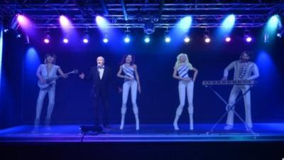 A man singing karaoke with a hologram featuring ABBA members