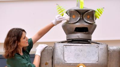 A large metal robot in the form of a person. It is taller than the lady cleaning its face. The robot has large lamps for eyes and yellow coiled ears.