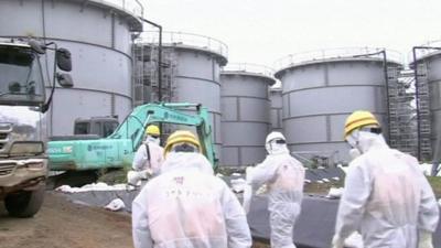 Workers at the plant wearing radiological protective suits, hard hats and masks walking towards large containers