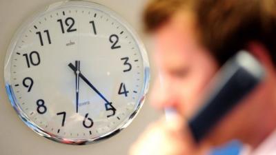 An office worker speaks on a telephone in front of a clock