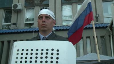 A man with a shield stands in front of a Russian flag